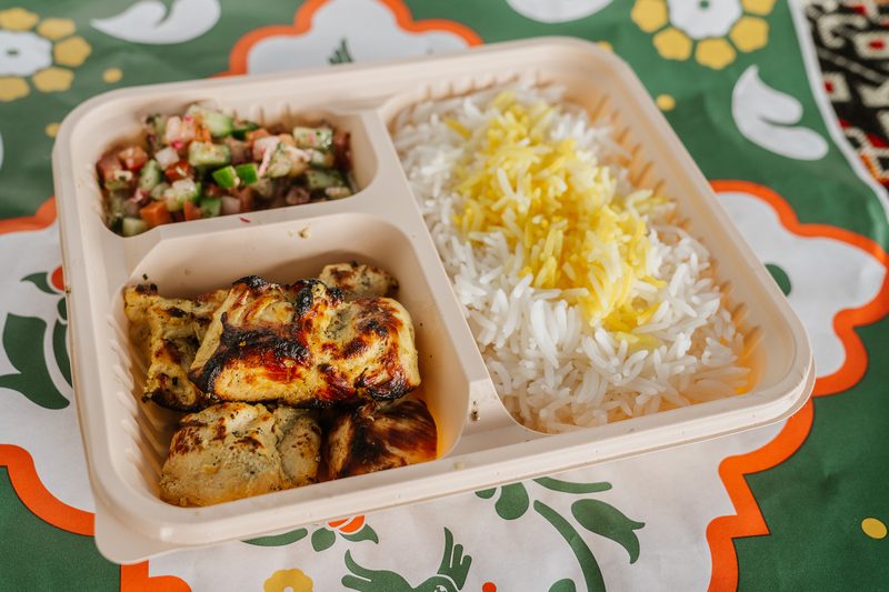 A chicken kabob meal in a three-compartment tray — grilled chicken, Shirazi salad and saffron rice — on KUKU patterned paper