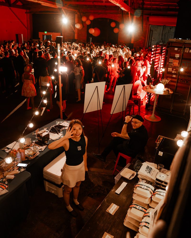 Seen from above: a KUKU team member at a counter with stacked meal boxes, a second team member seated nearby, and a crowd under string lights beyond
