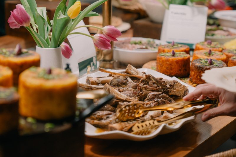 Braised lamb shanks served with gold tongs from a scalloped platter, with tahchin cakes and tulips along a catered table