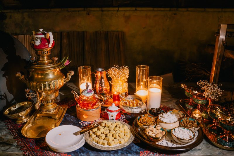 A samovar with tea glasses, a teapot, candles and plates of sweets and dried fruit on a patterned cloth