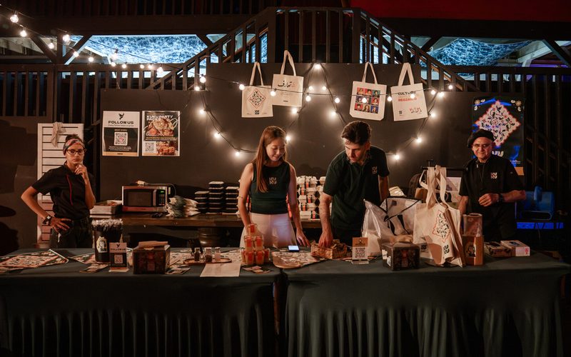 Four KUKU team members behind a long counter set with bags, boxes and signage, under string lights at an evening event